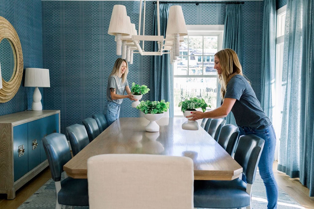 Two women in a formal dining room setting flowers on the table.